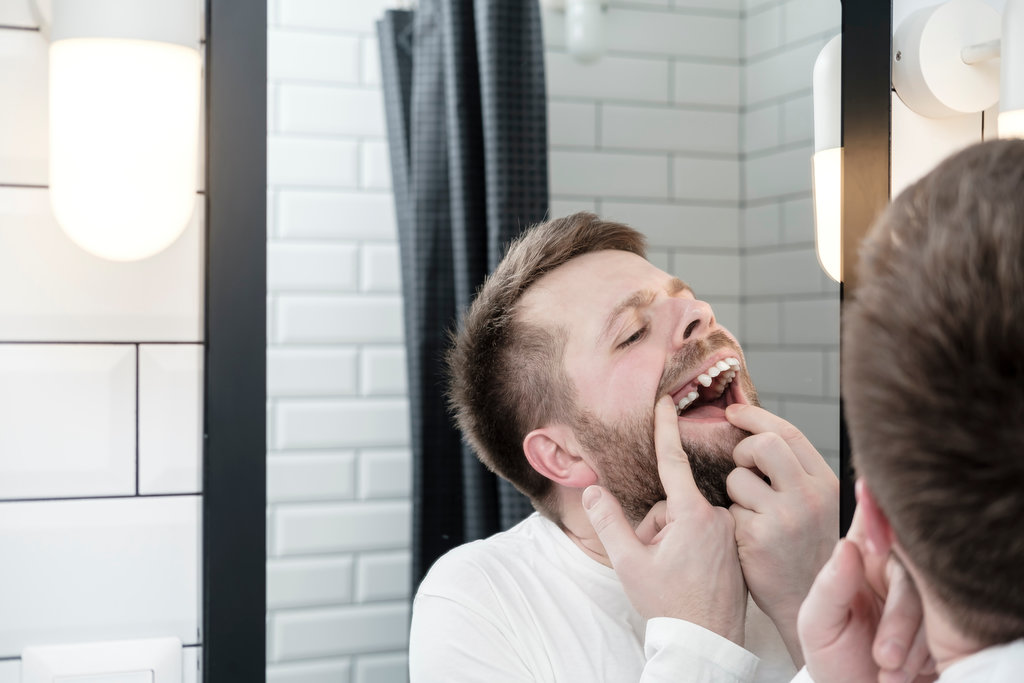 Upset young Caucasian man with a beard opened mouth and carefully examines the absence of a tooth, looking in the mirror in the bathroom.